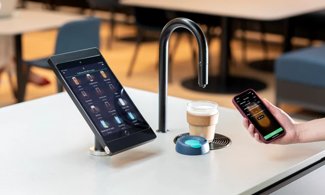 Image showing a matte black TopBrewer coffee machine with a reusable coffee cup under the swan neck and the TopBrewer app on an iPhone