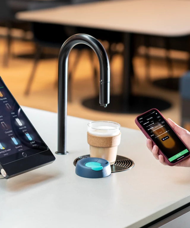 Image showing a matte black TopBrewer coffee machine with a reusable coffee cup under the swan neck and the TopBrewer app on an iPhone
