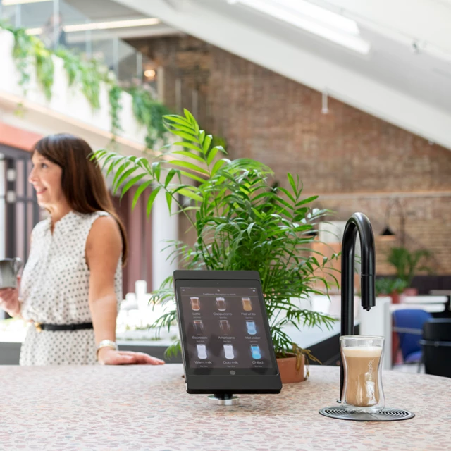Image showing a matte black TopBrewer commercial coffee machine in the foreground and two people chatting in the background