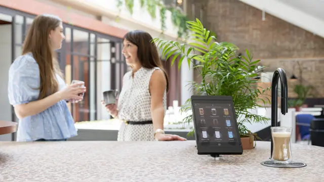 Image showing a matte black TopBrewer commercial coffee machine in the foreground and two people chatting in the background