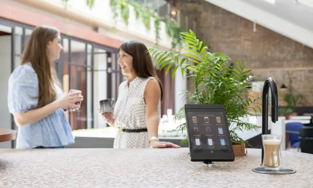 Image showing a matte black TopBrewer commercial coffee machine in the foreground and two people chatting in the background