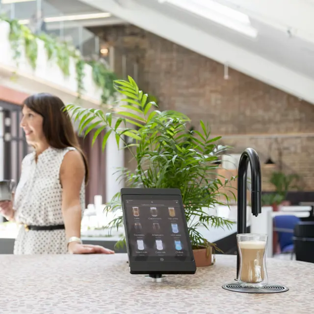 Image showing a matte black TopBrewer commercial coffee machine in the foreground and two people chatting in the background