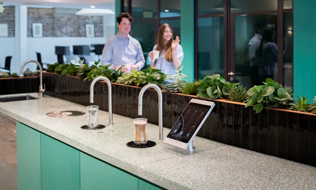 Image shows TopBrewer commercial coffee machine and TopWater built into a cabinet, with 2 people chatting in the background