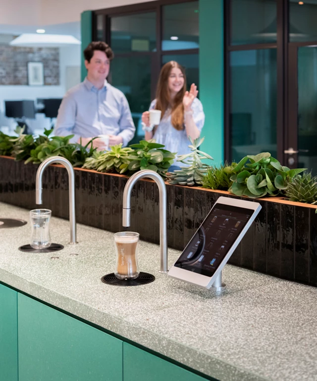 Image shows TopBrewer commercial coffee machine and TopWater built into a cabinet, with 2 people chatting in the background