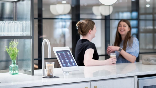 Image shows TopBrewer commercial coffee machine in the foreground with 2 colleagues chatting in the background