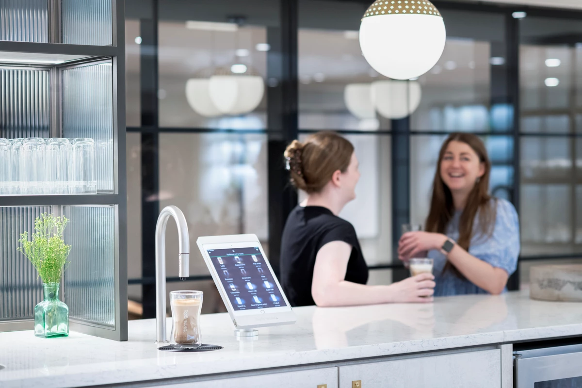 Image shows TopBrewer commercial coffee machine in the foreground with 2 colleagues chatting in the background